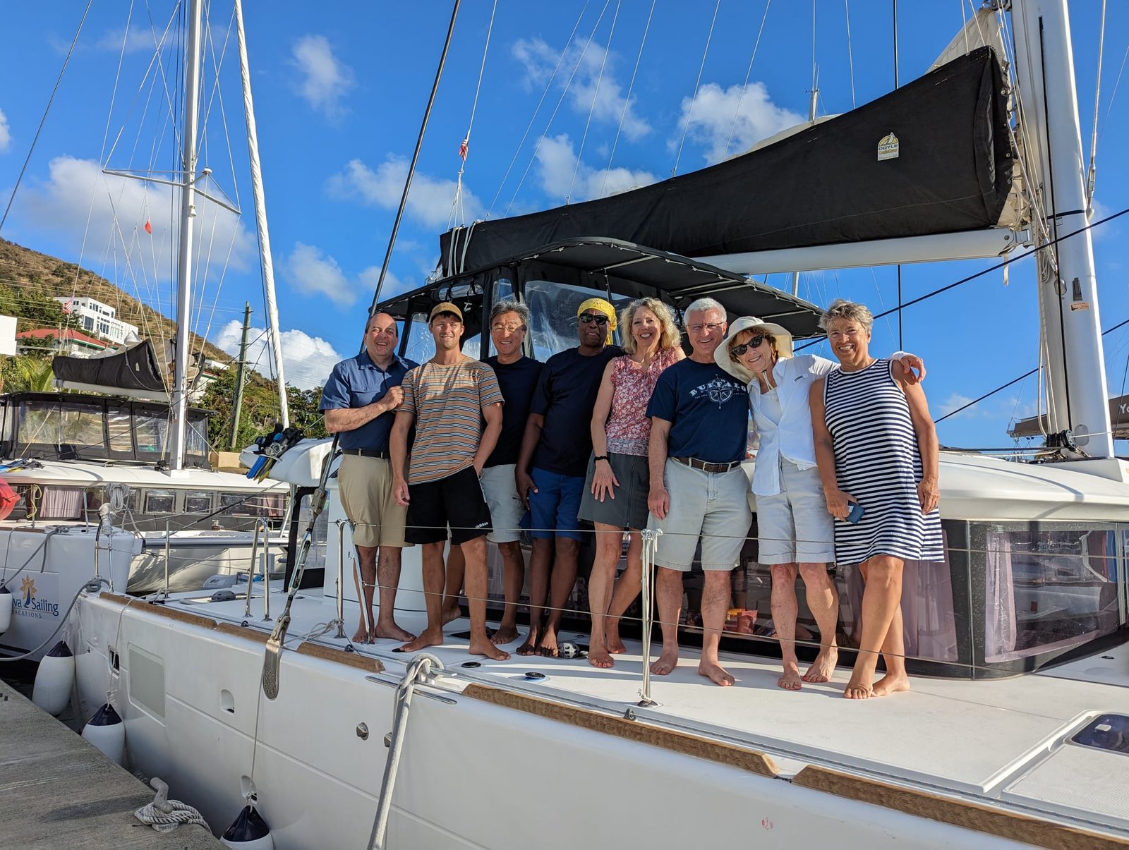 Group of friends on a yacht. A group of eight adults is standing and smiling on the deck of a docked white sailing catamaran on a sunny day. They are dressed in casual vacation attire, with masts and other boats visible in the background.