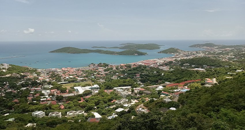 A high-angle, panoramic view of the coastal town of Charlotte Amalie, St. Thomas nestled among green hills, with a harbor filled with boats and small, rugged islands dotting the blue sea.